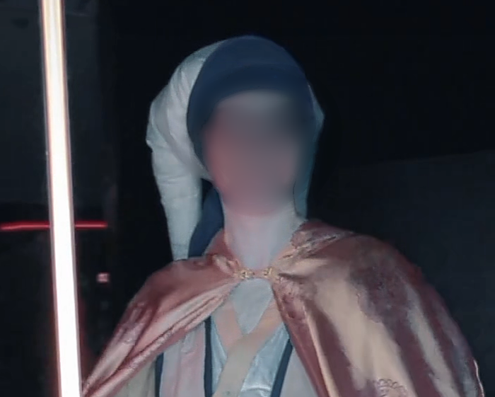 A picture of Chris in a Jedi cosplay. Chris's face is blurred. The cosplay has gold and blue thick shoulder straps (tabbards), a blue and gold belt, and white and tan tunic. Chris is wearing a headpiece that has two tail-like pieces hanging down past their shoulders that are designed as lekku from Twilek's from Star Wars. Chris is holding a black lightsaber that is set to yellow, almost gold, as the color.